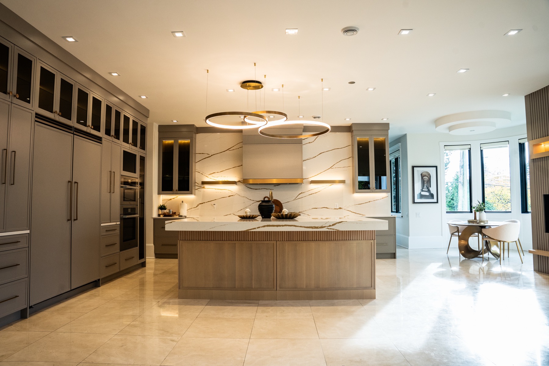 The same kitchen after renovation, with greige cabinetry, a fluted oak island, a book-matched stone backsplash and brass ring pendants